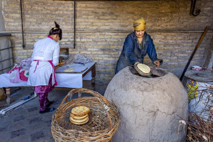 In a rustic bakery in Uzbekistan, two people prepare traditional bread. One person places dough in a clay oven while another works at a table. A basket with finished bread sits in the foreground, capturing the essence of Central Asian travel and culinary tradition.