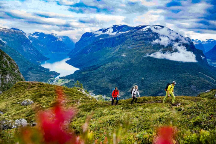 Three hikers walk along a grassy mountain trail in Norway, overlooking a serene lake surrounded by high mountain peaks under a cloudy sky.