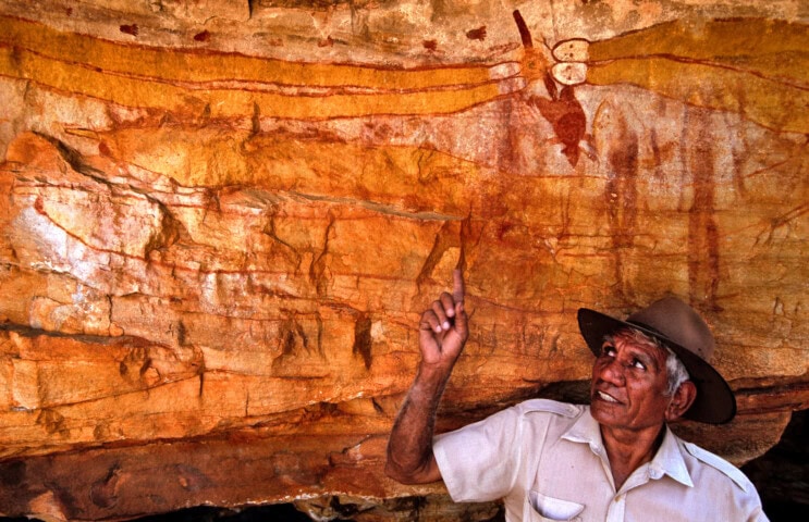 A man wearing a hat and a light-colored shirt points towards ancient Aboriginal rock art on a red, rocky wall in Australia.