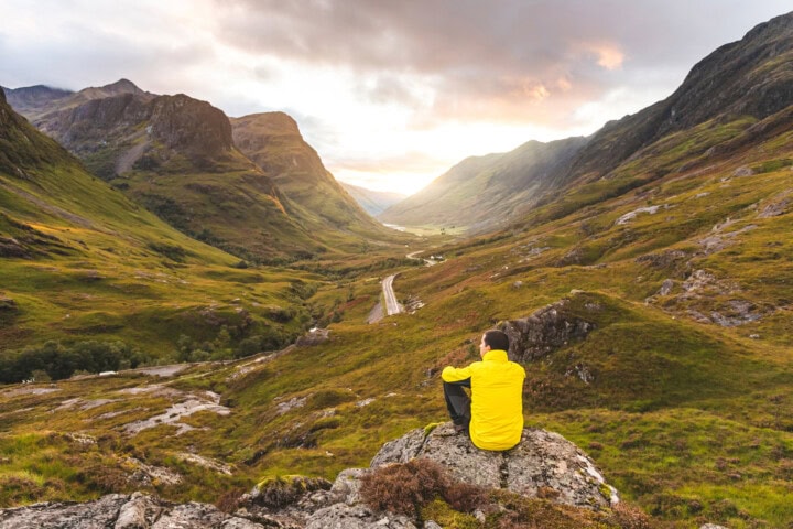 A person in a yellow jacket sits on a rocky outcrop overlooking a winding road in the lush, mountainous landscape of Scotland during sunset.