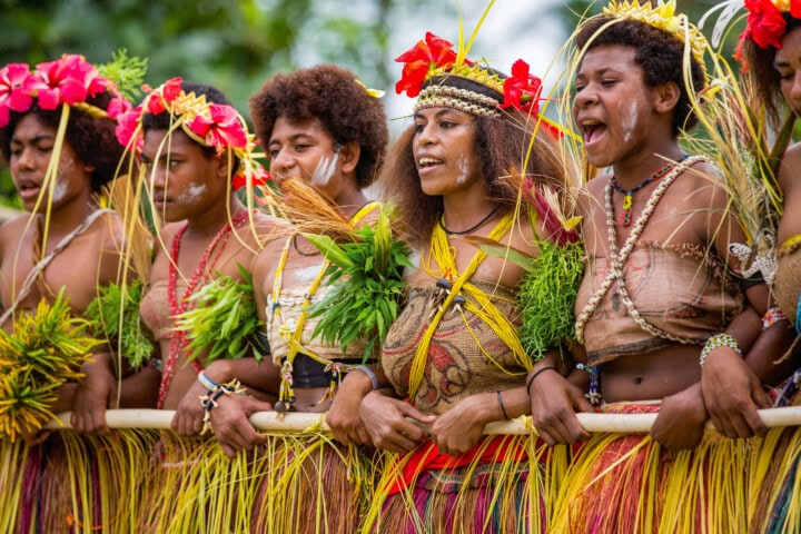 A group of women from Papua New Guinea adorned with traditional attire, including grass skirts and vibrant headdresses, sing and dance together outdoors.