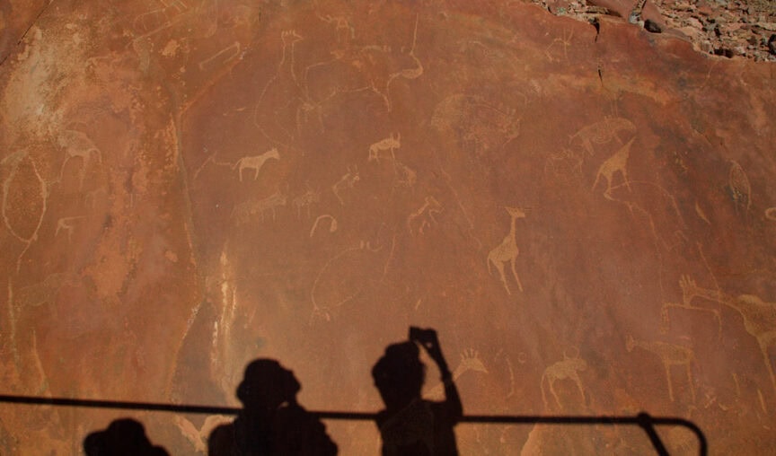 Shadows of people taking a photo of animal rock carvings on a large, weathered stone surface in Namibia, depicting various animals. This adventure unveils a wonderland of ancient artistry.