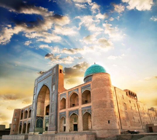 A historic Islamic architecture in Uzbekistan with a large arched entrance, intricate tile work, and a blue-domed roof, set against a backdrop of a vibrant sky with scattered clouds and sun rays.