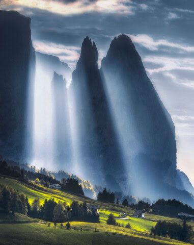Tall mountains with jagged peaks are illuminated by beams of sunlight breaking through clouds, casting light onto a green valley below. Small houses and trees are scattered in the foreground, creating a picturesque scene that invites tourism and travel enthusiasts to explore the beauty of Italy.
