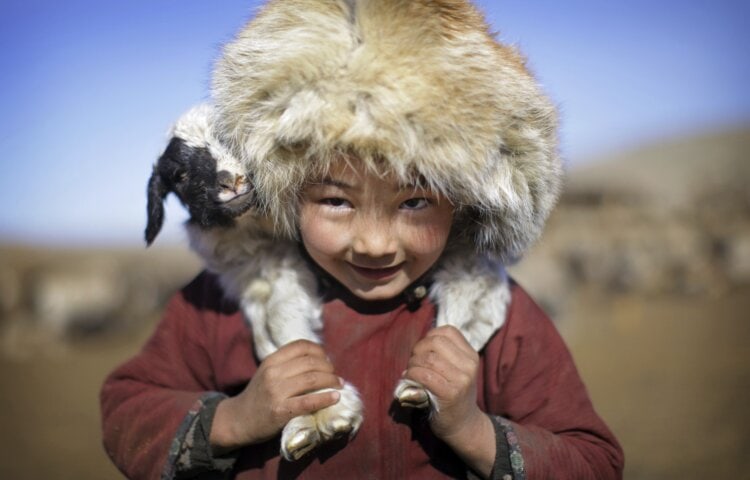 A child wearing a fur hat smiles while holding a small animal on their shoulders. The child is outdoors in Mongolia, with a blurred background of dry landscape.