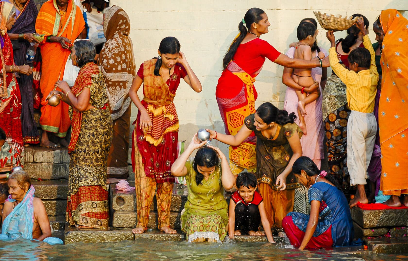 A group of people, including women and children, gather at a riverbank in Varanasi. One woman pours water over her head while others stand and interact. They're dressed in colorful traditional attire.