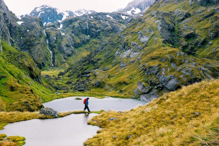 A person wearing a red jacket and backpack hikes along a grassy ridge by a small pond, surrounded by New Zealand mountains with patches of snow and waterfalls.