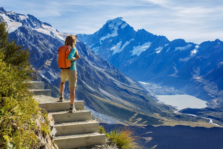 A person with an orange backpack stands on stone steps overlooking a mountainous landscape in New Zealand, with snow-capped peaks, valleys, and a distant lake.