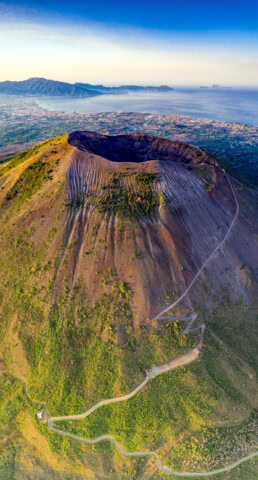 Aerial view of Mount Vesuvius showing its large volcanic crater, winding paths, and the city of Naples and coastline in the background.