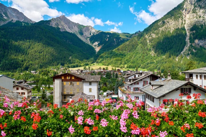 Colorful flowers in the foreground with a quaint Italian village and lush green mountains in the background under a blue sky with clouds.