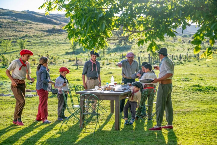 A group of people, including children and adults in traditional attire, gather around a table outdoors in a green, rural setting under a tree.