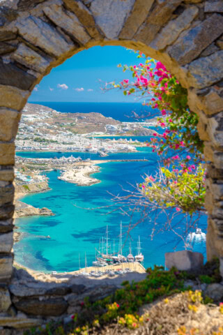 View of the Mykonos coastline in Greece, with blue waters, sailboats, and white buildings, framed by a stone arch with flowering pink bougainvillea.