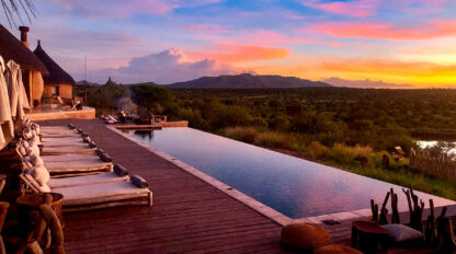 A luxurious infinity pool on a wooden deck overlooks a lush green landscape and distant mountains under a vibrant sunset sky. Reserved lounge chairs and a fire pit are visible to the left, perfect for those exploring Namibia during the stunning Green Season.