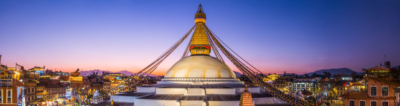 Panoramic view of the Boudhanath Stupa in Kathmandu, Nepal at dusk, with prayer flags and city buildings visible under a purple sky.