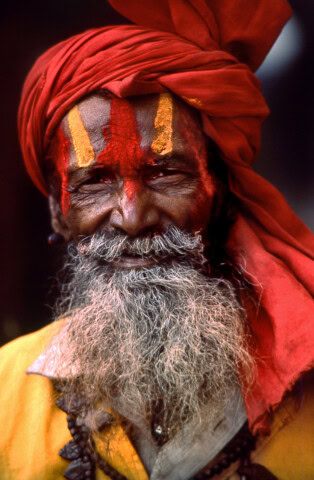A man with a long gray beard and a red turban smiles. He has red and yellow paint marks on his forehead and wears a necklace with dark beads, evoking the rich cultural traditions of Nepal.