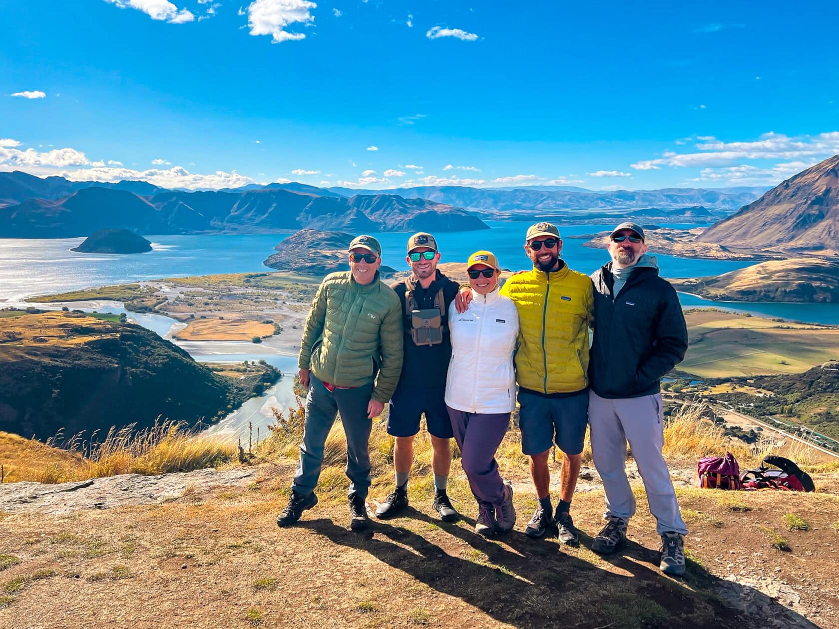 Five people wearing outdoor clothing stand on a hilltop, smiling at the camera, with a scenic view of lakes and mountains in the background.