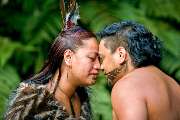 Two individuals with traditional tattoos and feathers touch foreheads in an intimate gesture, set against the lush greenery of New Zealand.