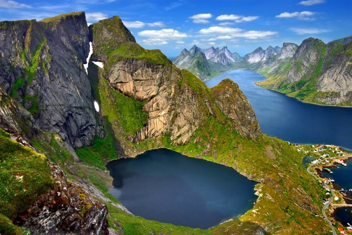 Aerial view of Reinebringen in Lofoten, Norway. It features steep, rocky mountains, two lakes, coastal fjords, and a small village with blue skies and scattered clouds above.