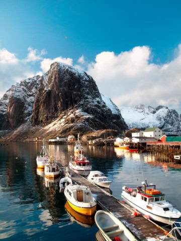 A small harbor in Norway with several boats docked, set against a backdrop of snowy mountains and calm water under a partly cloudy sky, offers a picturesque travel destination for tourism enthusiasts.