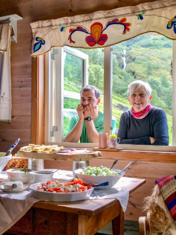 Two people smile while sitting by an open window of a wooden cabin in Norway, with mountains and a waterfall in the background. A table in the foreground displays various dishes of food.