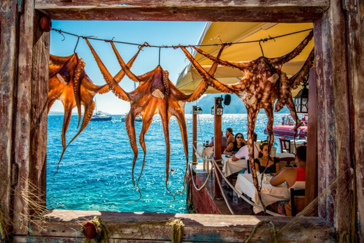 Octopuses hang to dry in the sun in an open wooden frame overlooking a seaside restaurant in Greece, with patrons and boats in the background.
