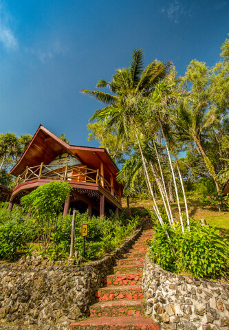 A wooden house with a sloped roof and a wrap-around porch stands elevated with red brick steps leading up. Palm trees and lush greenery surround the house under a clear blue sky, creating an idyllic travel destination reminiscent of Palau's scenic tourism spots.