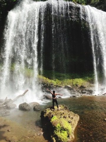 A person stands with arms outstretched on a rock in front of a large, cascading waterfall, surrounded by lush greenery in Palau's tropical paradise—a true travel destination dream.