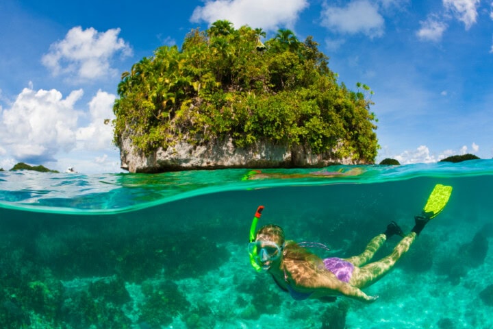 A person snorkeling in clear blue water near a small, lush island under a bright sky with scattered clouds in Palau.