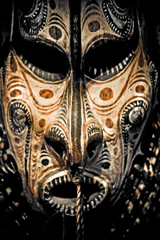 Close-up of an intricately designed tribal mask with dark eye holes, detailed geometric patterns, and a rope hanging from the mouth. The mask, reminiscent of Papua New Guinea craftsmanship, features earthy tones and conveys a traditional aesthetic.