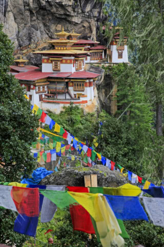 Colorful prayer flags lead to the Taktshang Monastery, also known as Tiger's Nest, perched on a cliffside amidst lush greenery and rocky surroundings in Bhutan.