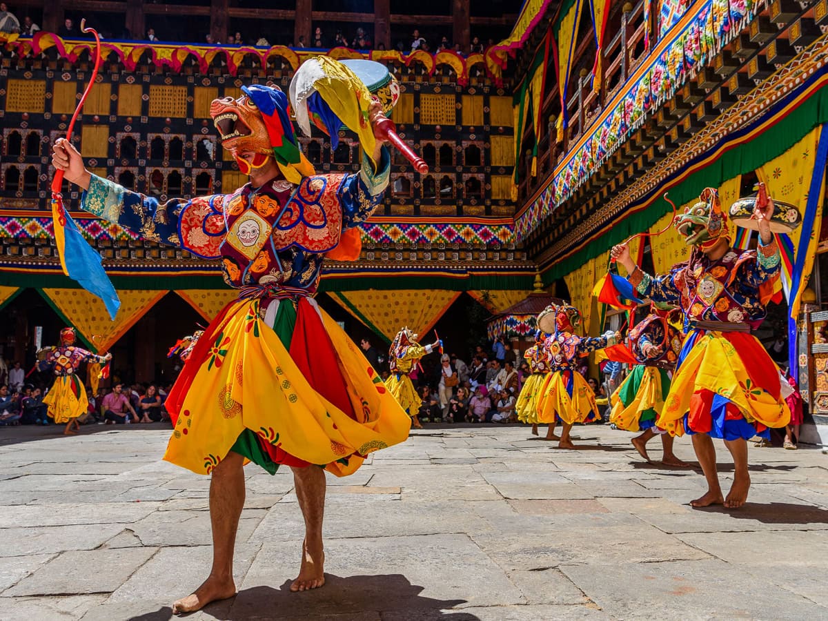 In a vibrant courtyard, colorfully dressed dancers donning masks perform a traditional dance as part of Bhutan's renowned Tshechu festival, captivating an eager audience with their rhythmic movements and vivid costumes.