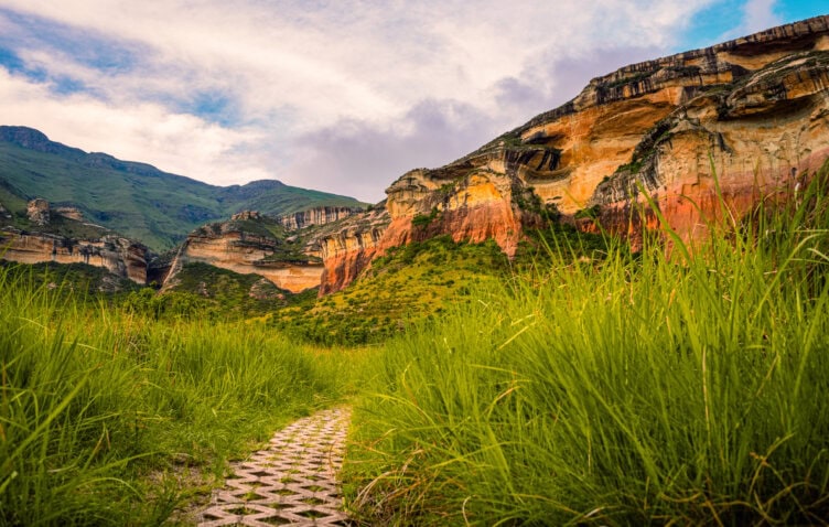 A paved path winds through tall grass toward colorful, layered sandstone cliffs and green hills under a partly cloudy sky in one of the best parks South Africa has to offer.