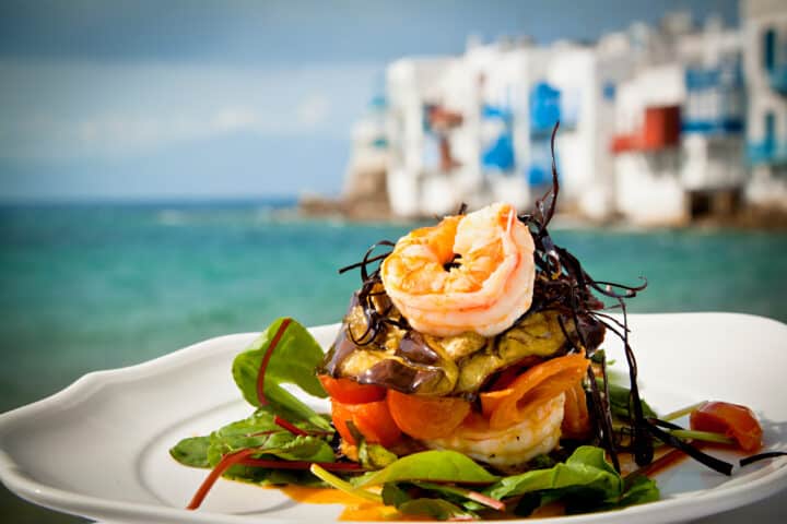 A gourmet seafood dish with prawns, vegetables, and greens on a white plate is set against a coastal Greek backdrop featuring white buildings with blue accents.