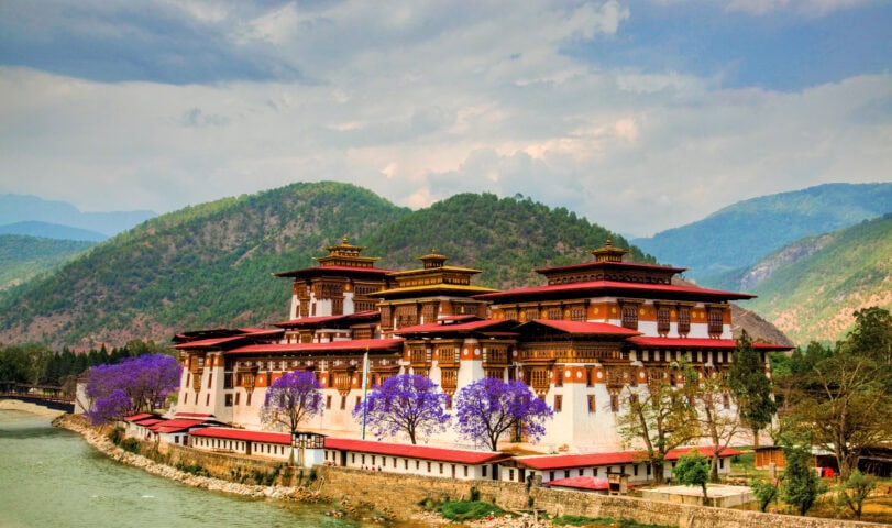 A large traditional Himalayan fortress with red, white, and gold architecture sits beside a river, surrounded by purple-flowered trees and forested mountains under a cloudy sky.