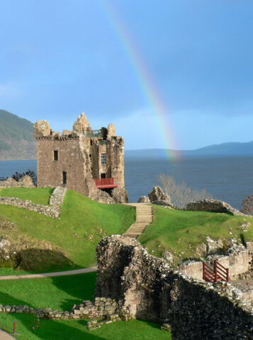 The stone ruins of a castle overlook a large body of water with a rainbow arching in the sky, making it a perfect travel destination in Scotland. Green grass carpets the surrounding landscape, enhancing its picturesque charm and ideal for tourism.