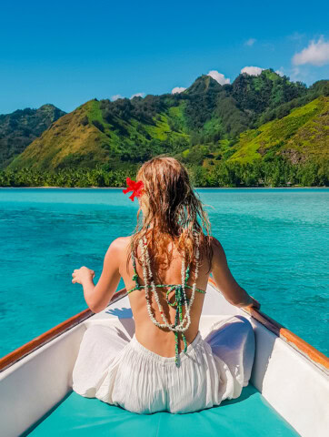 A woman with a flower in her hair and wearing a beaded necklace sits on a boat facing the turquoise waters of French Polynesia, with green mountains in the background.