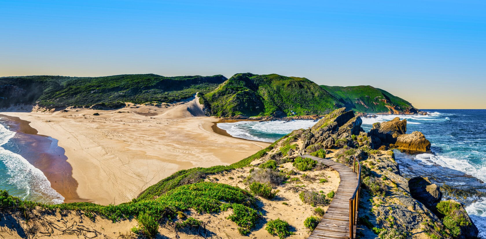 A sandy beach with waves on both sides, a wooden walkway in the foreground, and green hills in the background under a clear blue sky—one of the best parks South Africa has to offer.