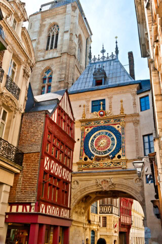 Historic clock mounted on an archway with intricate detailing, surrounded by old buildings and a tall church tower in the background, making it a must-see for anyone interested in travel and tourism in France.