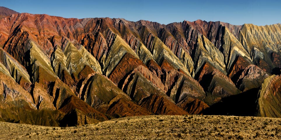 A panoramic view of multicolored, zigzag-patterned hills in Argentina under a clear blue sky, with red, orange, yellow, and brown hues distinctly visible. The foreground is a barren, grassy area.