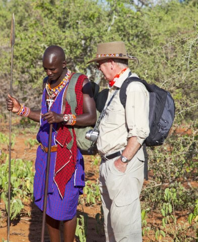 Two men stand outdoors. One wears traditional Maasai attire and holds a spear; the other, on a tour, wears a hat and carries a camera. They are surrounded by sparse vegetation.