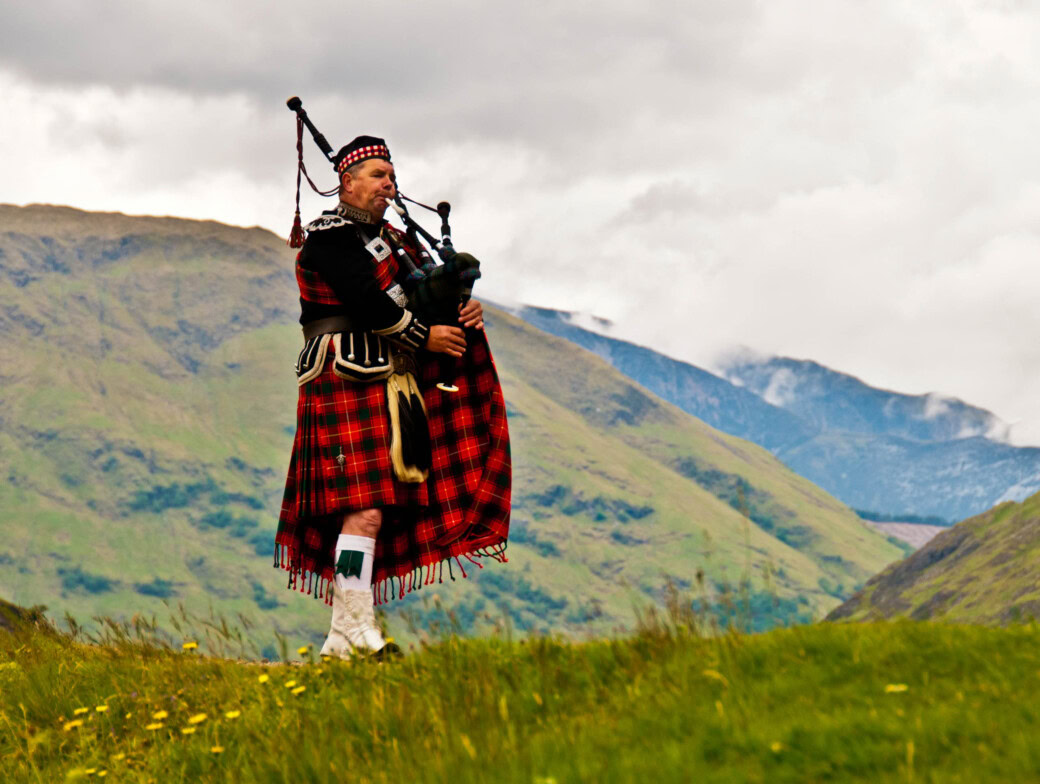 A piper plays the bagpipes on a hillside in traditional Scottish dress including kilt.