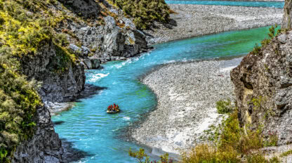 A group of people whitewater rafting down a winding turquoise river in Queenstown, surrounded by rocky cliffs and greenery, is one of the top attractions and thrilling things to do.