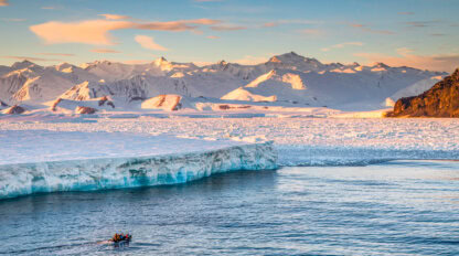 A small boat with people approaches a large ice shelf on calm water during an Antarctica tour, with snow-covered mountains and a colorful sky in the background.