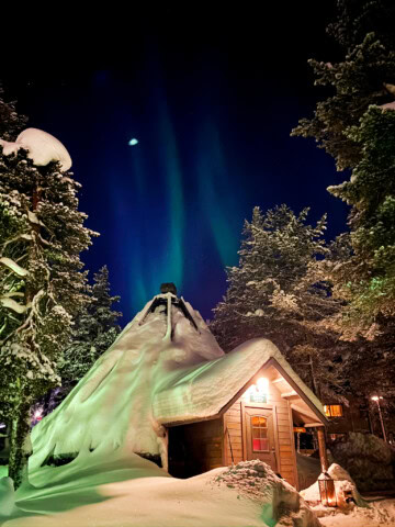 A snow-covered cabin and teepee under a night sky illuminated by the Northern Lights, with surrounding snow-laden trees in the tranquil wilderness of Finland.