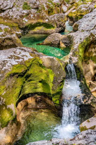 A small waterfall cascades into a series of clear turquoise pools surrounded by moss-covered rocks in the enchanting natural landscape of Slovenia.