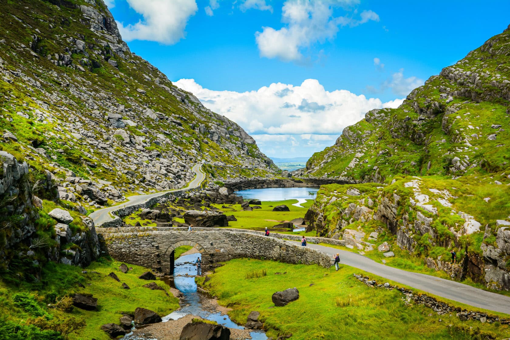 Explore Ireland's best region with a scenic winding road through a lush green valley, rocky hills, and a stone bridge over a stream. A small lake completes this picturesque setting under a partly cloudy blue sky, beckoning visitors to indulge in its serene beauty.