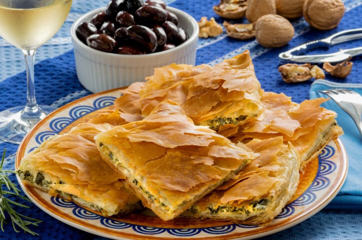 A plate of phyllo pastry triangles filled with spinach and cheese, reminiscent of Greece, accompanied by a bowl of black olives and a glass of white wine on a blue tablecloth with walnuts nearby.