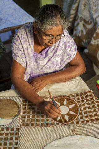 An older woman with glasses and grey hair, wearing a purple patterned dress, paints a detailed design on a large fabric in a Sri Lankan workshop, capturing the essence of local craftsmanship that attracts tourism and travel enthusiasts from around the world.