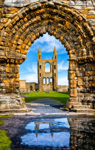 Ruins of an ancient stone cathedral with two towers stand in the distance, framed by a large arched doorway with intricate brickwork. A puddle in the foreground reflects parts of the structure, capturing a timeless scene reminiscent of Scotland's historic charm.