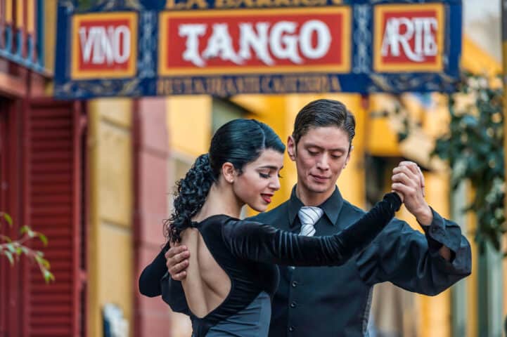 A couple dressed in black dance the tango on a street, with a colorful "Tango" sign in the background, evoking the passionate spirit of Argentina.